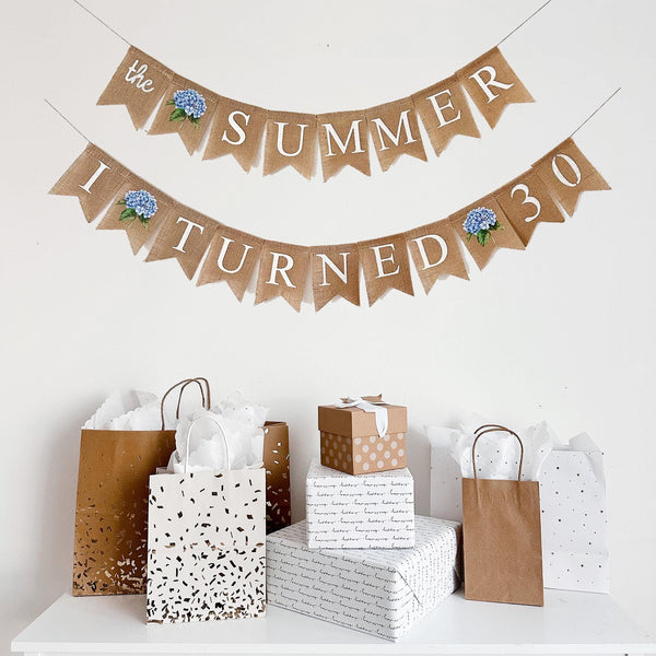 a banner that reads "The SUMMER I TURNED 30" with a blue flower design, and there are gift bags and wrapped presents underneath it.