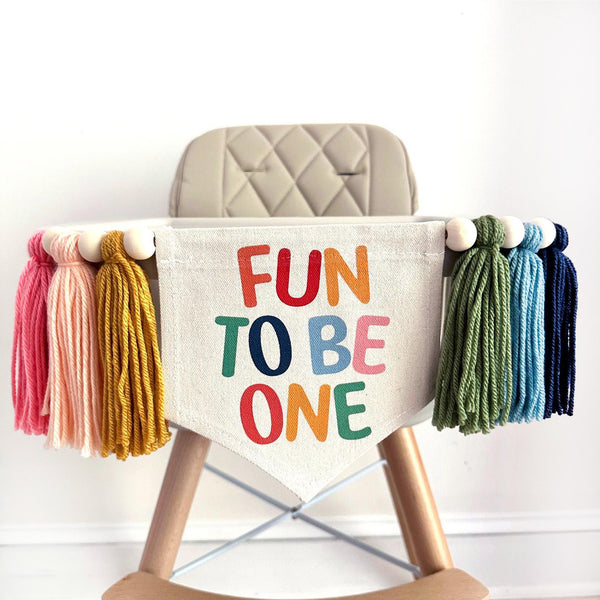 A first birthday high chair banner with colorful tassels on either side of a canvas flag with the words "FUN TO BE ONE" printed on it in multi colors.
