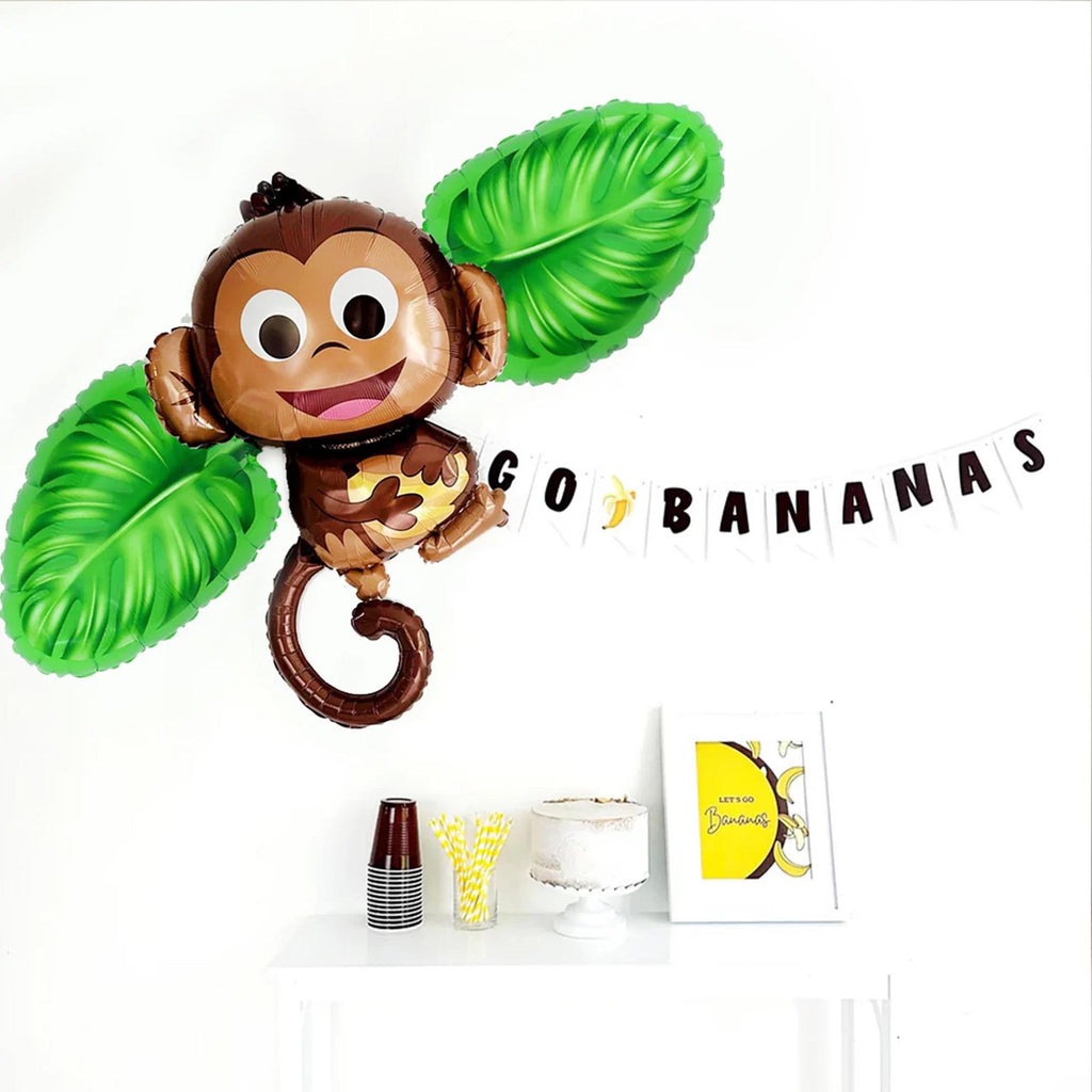 A monkey balloon with a banner that says "GO BANANAS" hanging on a wall. Below the monkey balloon, there is a table with a cake, cups, and a book.