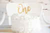 A white cake with a gold glitter One cake topper on it.