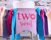 A banner with the text "two sweet" and a pink doughnut graphic, surrounded by colorful ribbons and tassels.