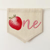 A beige fabric banner with a red strawberry and the word "one" written on it.