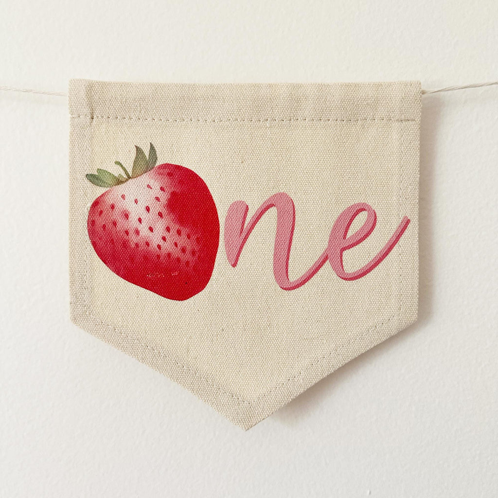 A beige fabric banner with a red strawberry and the word "one" written on it.