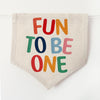 A beige fabric banner with the text "FUN TO BE ONE" printed on it in a playful, colorful font.