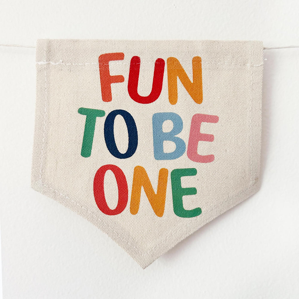 A beige fabric banner with the text "FUN TO BE ONE" printed on it in a playful, colorful font.