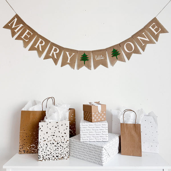 a festive scene with a "Merry Little" banner hanging above a display of wrapped gifts and brown paper shopping bags.
