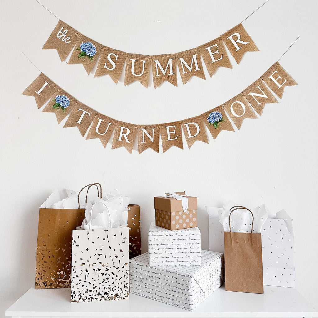 a banner that says "The SUMMER I TURNED ONE" and a collection of gift bags and boxes on a white surface.
