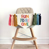 A beige high chair with a colorful banner that says "FUN TO BE ONE" with matching colorful tassels on either side.