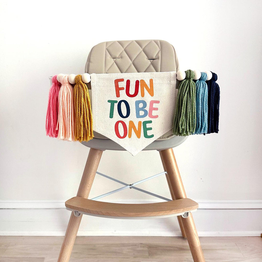 A beige high chair with a colorful banner that says "FUN TO BE ONE" with matching colorful tassels on either side.