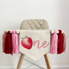 A beige high chair with a white banner featuring a red strawberry and the number "one" on it, along with tassels in pink and red.