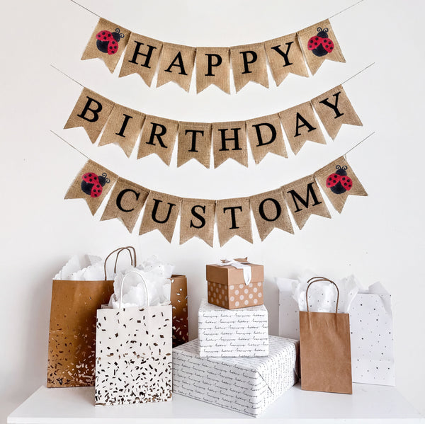 A happy birthday burlap banner strung on three strings. The happy is at the top, birthday second, and custom on the third row. Hand painted red & black ladybugs are at the beginning and end of the word happy and custom.