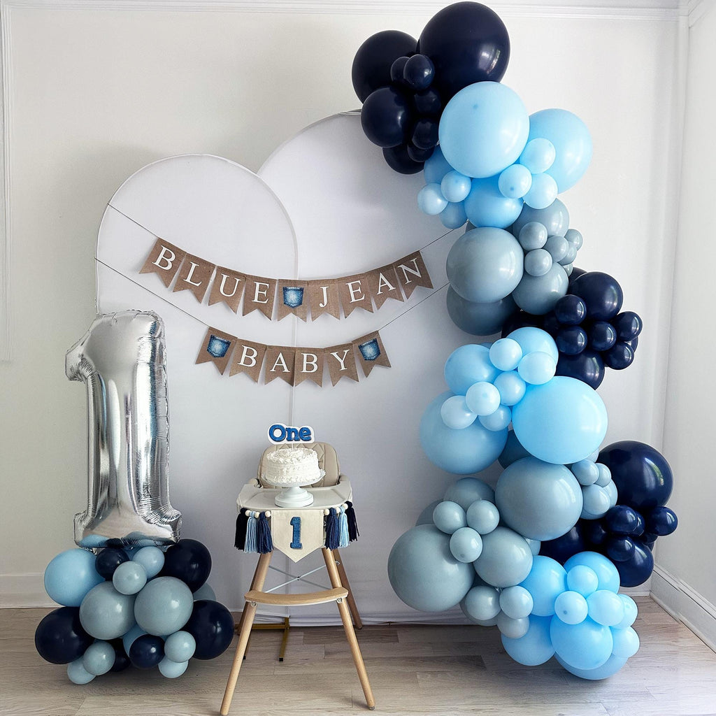 a room decorated for a baby's first birthday celebration. The centerpiece is a large balloon arch in shades of blue and gray, with a banner that reads "Blue Jean Baby". A cake is placed on a table, and a silver number one balloon is leaning against the wall. The room has a white wall and a wooden floor, creating a clean and festive atmosphere.