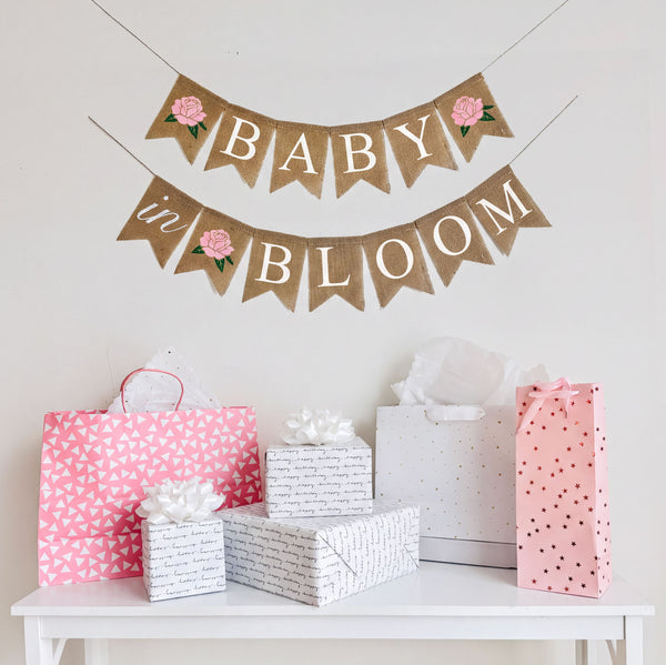 A Baby in Bloom baby shower banner in white letters on burlap flags. Starting with a pink hand stenciled rose, the first line says baby with a pink flower after the word, the second line reads in bloom with a pink rose in between the words.