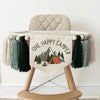A beige high chair with a banner that says "ONE HAPPY CAMPER" and features an illustration of a tent and mountains.