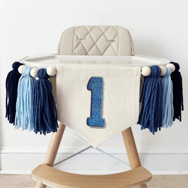 This image shows a high chair decorated with dark blue and light blue tassels and a banner that displays the number 1 in a denim print.
