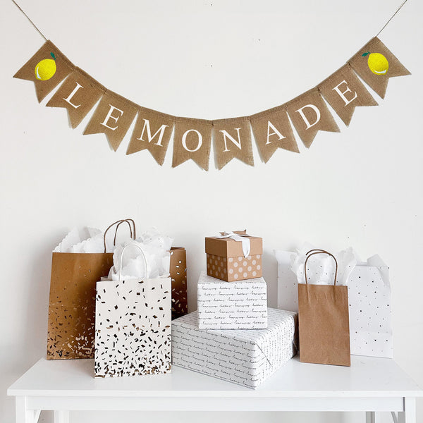 a white table with various gift bags and boxes on it, and a banner with the word "LEMONADE" hanging above.