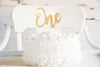 A white cake with a gold glitter One cake topper on it.
