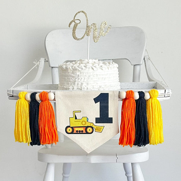 A 6  x 7 inch canvas center flag with a black number 1 in the bucket of a bulldozer.  On each side there is a 5 inch orange, black and yellow yarn tassel each separated by a wooden bead. It is hung on a high chair in the photo.