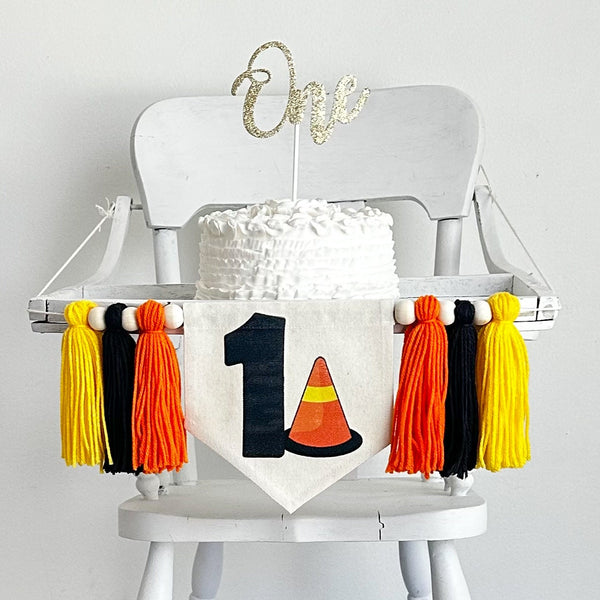 A 6  x 7 inch canvas center flag with a black number 1 and an orange caution cone beside it. On each side there is a 5 inch orange, black and yellow yarn tassel each separated by a wooden bead. It is hung on a high chair in the photo.