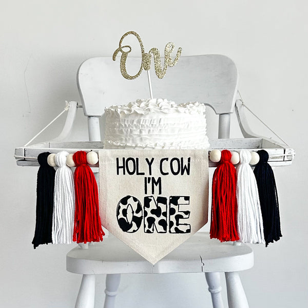 A 6 x 7 inch canvas center flag with the words HOLY COW I&#39;M in black and the word ONE in a cow print pattern. There is a 5 inch red, white and black yarn tassel on each side each separated by a wooden bead for a total of 6 tassels.