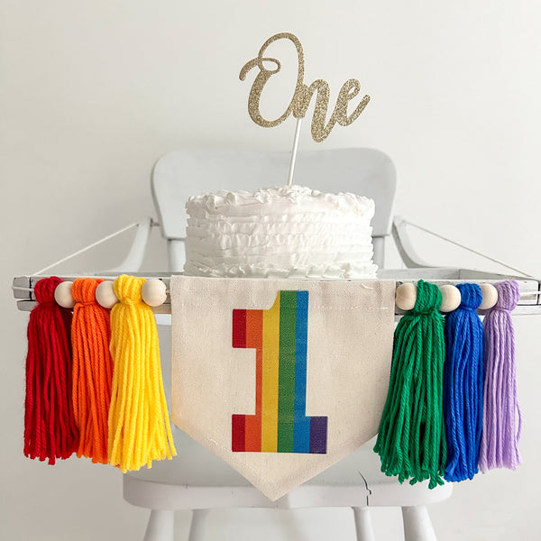 A 5 by 7 inch canvas center flag with a rainbow number 1. There is a 5 inch red, orange, yellow, green, blue and lavender colored yarn tassel on each side each separated by a wooden bead for a total of 6 tassels.