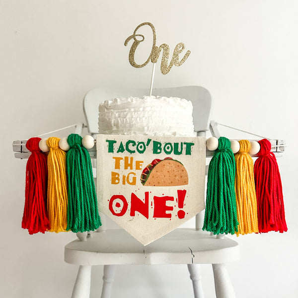 A 5 by 7 inch canvas center flag with a taco image and the words TACO BOUT THE BIG ONE in green, gold and red. There is a 5 inch green, gold and red colored yarn tassel on each side each separated by a wooden bead for a total of 6 tassels.