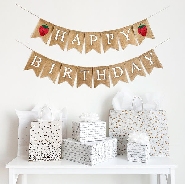 A happy birthday burlap banner strung on two strings. The happy is at the top and the birthday below.  Hand painted strawberries are at the beginning and end of the word happy.  The strawberries are painted in red with a green stem and black seeds.
