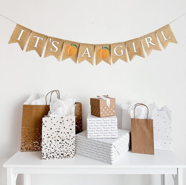 A burlap banner made of fishtail shaped flags with the words It&#39;s A Girl in white letters. There is an image of a peach between each word for a total of two peaches.