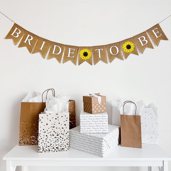 Burlap Bride to Be banner with sunflowers. Written in white letters on burlap flags, this banner reads, bride to be with a vibrant hand stenciled sunflower between the words bride to be.