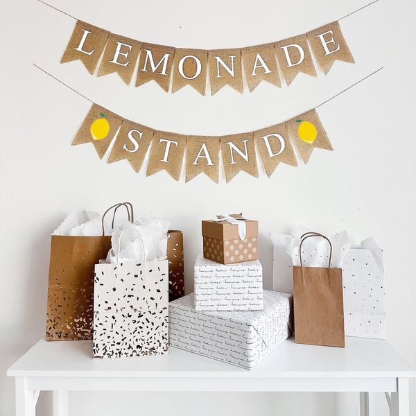 This is a burlap banner made from 4.5 by 6.5 inch fishtail shaped flags. Each flag has a white letter. The word LEMONADE is on the top string. The word STAND is on the bottom string with an image of a lemon on either end.