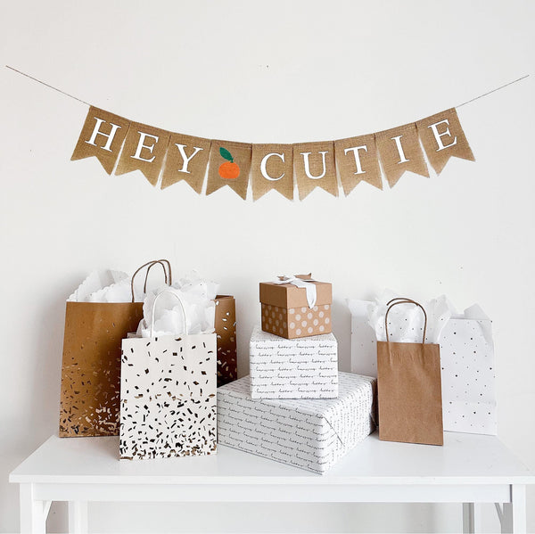 This is a burlap banner made from fishtail shaped flags. Each flag measures 4.5 by 6.5 inches and has one letter or image on it. HEY CUTIE is spelled out in white letters. There is an image of a tangerine in the middle of the words.
