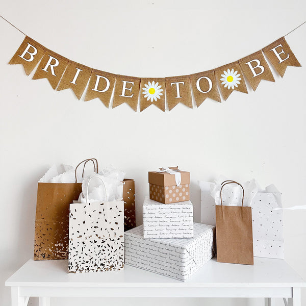 Beige burlap banner with bride to be written out on each flag in white letters. White hand stenciled daisy flags hand between each word. Perfect for a rustic bridal shower or engagement celebration.