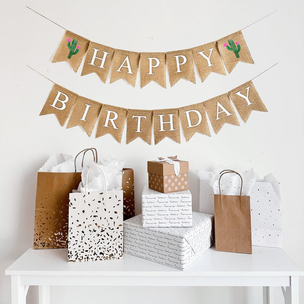 A happy birthday burlap banner strung on two strings. The happy is at the top and the birthday below.  Hand painted cacti with pink flowers are at the beginning and end of the word happy.  The cacti are painted in green with pink flowers.
