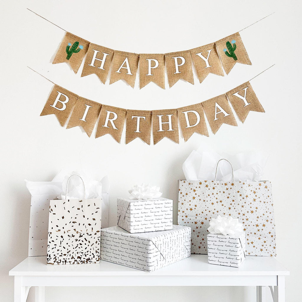 A happy birthday burlap banner strung on two strings. The happy is at the top and the birthday below.  Hand painted cacti with blue flowers are at the beginning and end of the word happy.  The cacti are painted in green with blue flowers.