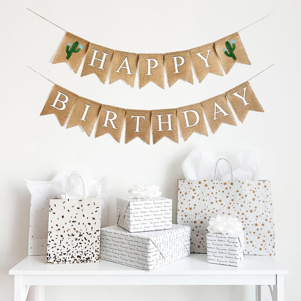 A happy birthday burlap banner strung on two strings. The happy is at the top and the birthday below.  Hand painted cacti with blue flowers are at the beginning and end of the word happy.  The cacti are painted in green with blue flowers.