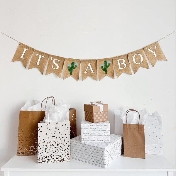 An It’s a Boy burlap banner strung on one string. Hand painted cacti are between the word it’s and a, and then between the words a and boy.  The cacti are painted in green with light blue flowers.