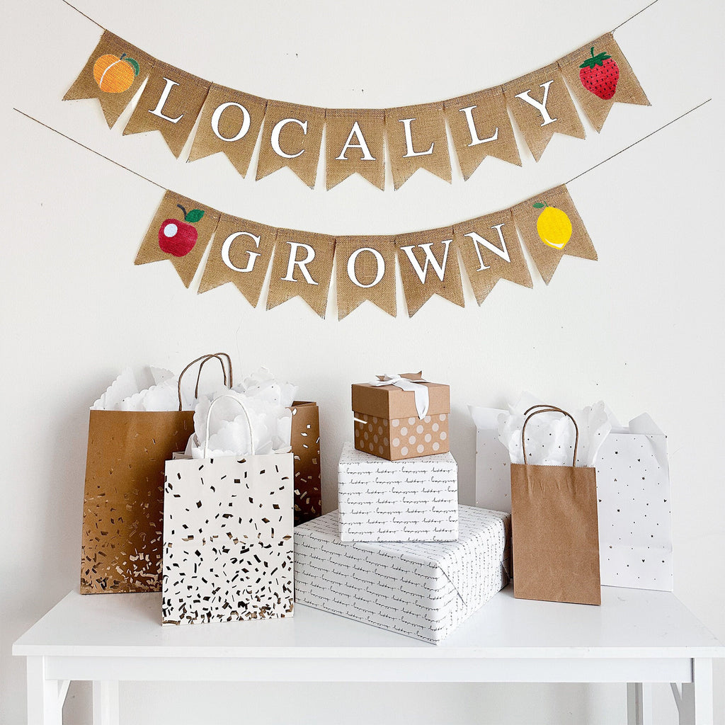 This is a burlap banner of fishtail shaped flags with the word Locally on the top string with a peach on the left side and a strawberry on the right side. The bottom string features the word Grown with an apple on the left and a lemon on the right.