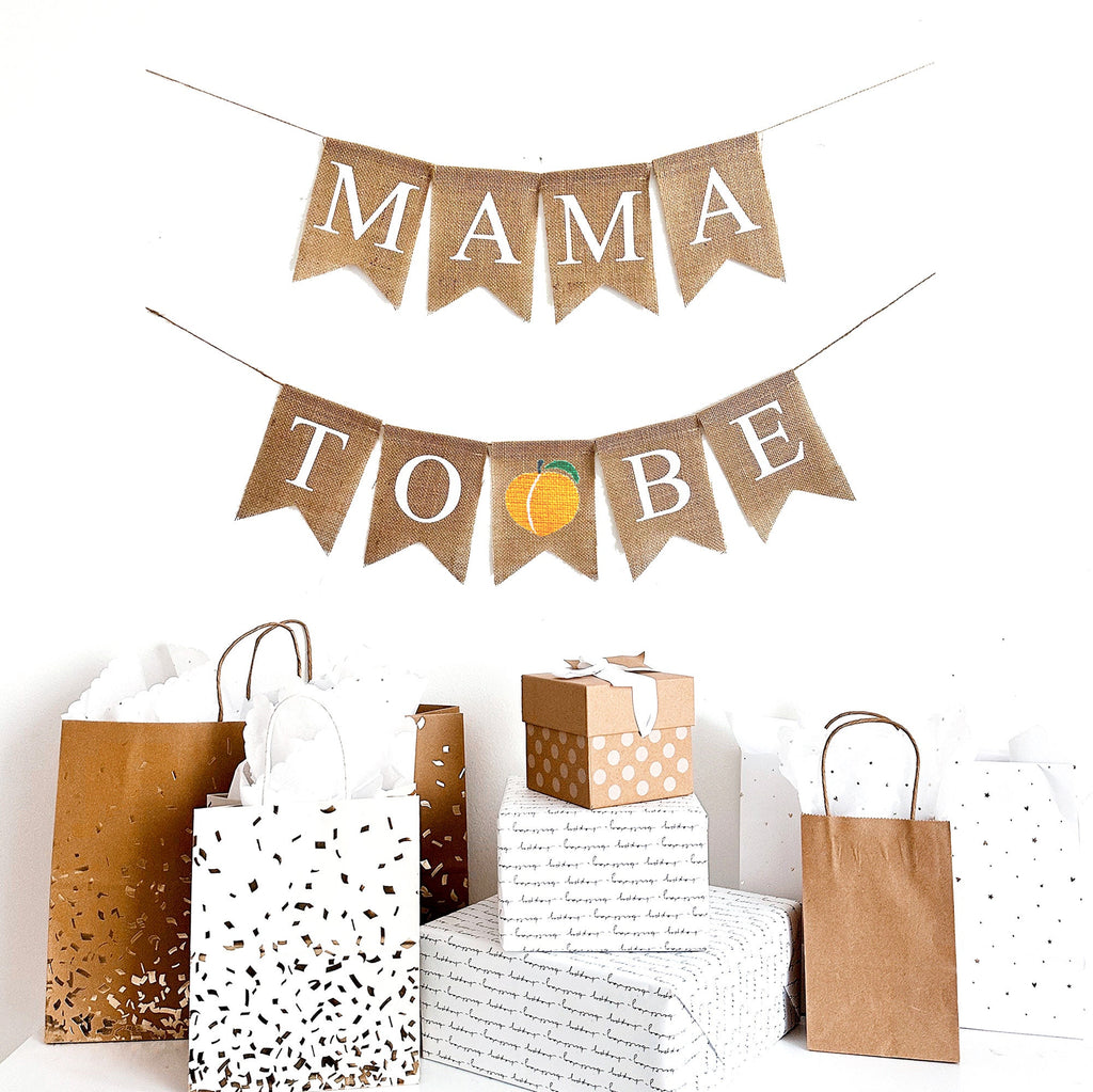 This is a Mama to Be burlap banner on fishtail shaped flags. Mama to Be is in white capital letters on two strings. The top string says Mama and a hand painted peach is in between the words to be on the bottom string.