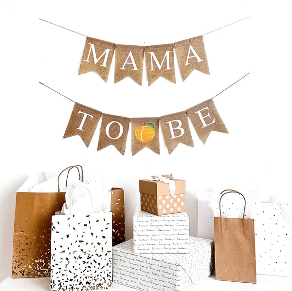 This is a Mama to Be burlap banner on fishtail shaped flags. Mama to Be is in white capital letters on two strings. The top string says Mama and a hand painted peach is in between the words to be on the bottom string.