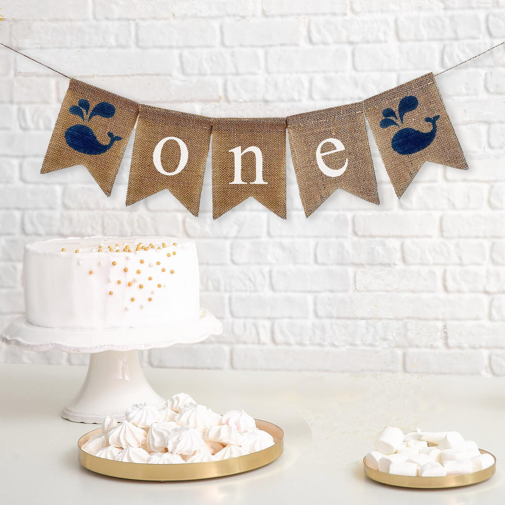 This is a burlap banner made of 5 fishtail shaped flags. The word ONE is spelled out in white lowercase letters on the middle three flags. There is an image of a navy whale on each end of the banner. The flags measure 4.5 by 6.5 inches each.