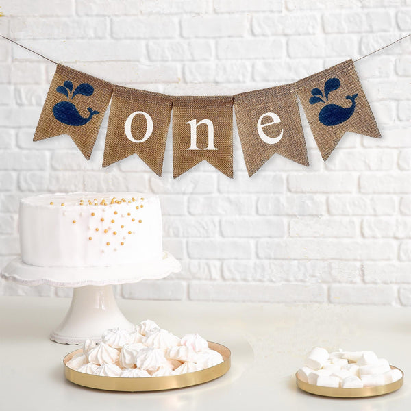 This is a burlap banner made of 5 fishtail shaped flags. The word ONE is spelled out in white lowercase letters on the middle three flags. There is an image of a navy whale on each end of the banner. The flags measure 4.5 by 6.5 inches each.
