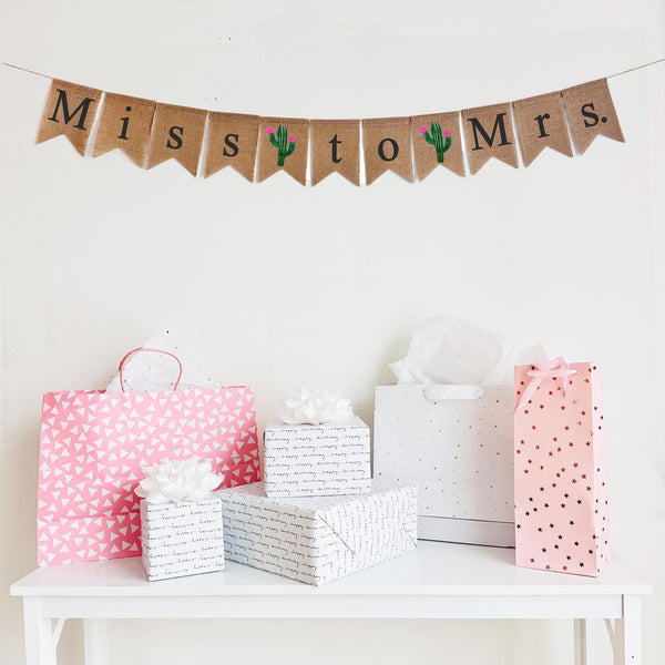 Miss to Mrs bridal shower burlap banner features flags that spell out the words Miss to Mrs with cactus flags between each word. Includes 2 image flags. The letters are painted in black. The cactus is green with pink flowers.