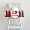 This is a banner that can hang on a highchair with a canvas center flag with the words MY FIRST RODEO in red letters with a cowboy hat at the top. There is a red, tan and brown yarn tassel hanging down on either side of the center banner.