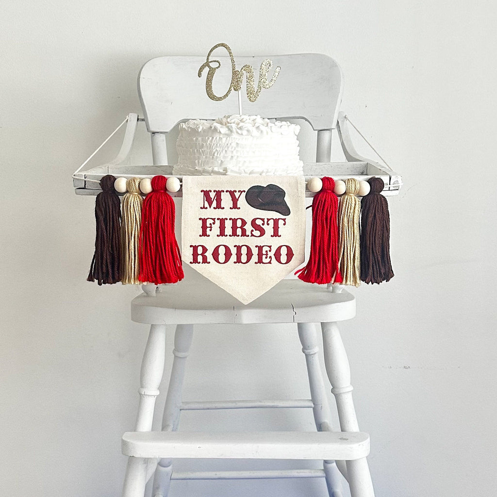 This is a banner that can hang on a highchair with a canvas center flag with the words MY FIRST RODEO in red letters with a cowboy hat at the top. There is a red, tan and brown yarn tassel hanging down on either side of the center banner.