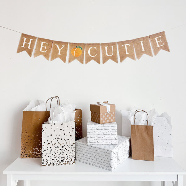 This is a burlap banner with fishtail shaped flags on one string. The words Hey Cutie are stenciled in white, and there is an image of a peach in between the words. There is one letter on each flag. The flags measure 4.5&quot; by 6.5&quot; each.