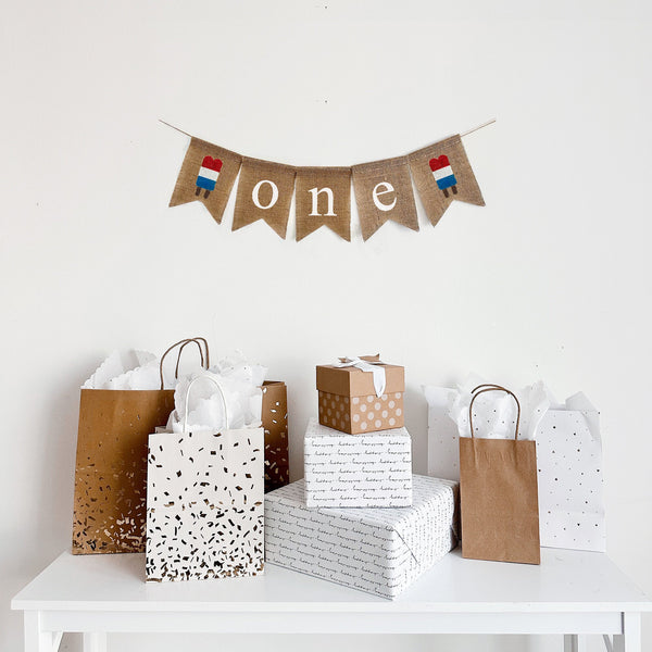 A burlap banner that reads O N E with a red white and blue popsicle on each end. Perfect for a 4th of July First Birthday celebration.