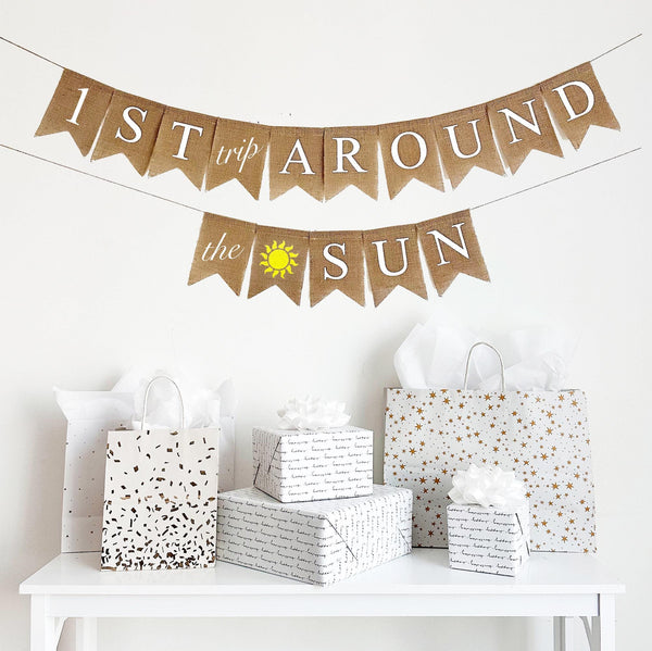A two tiered burlap banner. First line reads first trip around, and the second line reads the sun with a yellow sunshine image.