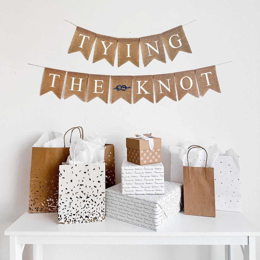 This is a burlap banner made of fishtail shaped flags that measure 4.5 by 6.5 inches each. The top string says TYING in white letters. The bottom string says THE KNOT in white letters with an image of a rope knot in navy in between the words.