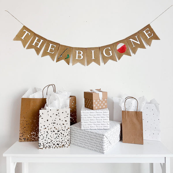 A burlap banner that reads the big one with a fishing rod with a green fish on the line between the and big and a red and white bobber replacing the o in one.