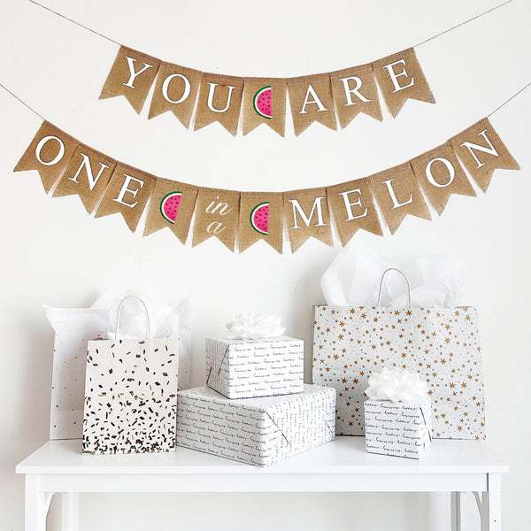 A two tiered burlap banner. The first tier reads you are with a pink & green watermelon between the words. The second tier reads one in a melon with the same watermelons separating the words.